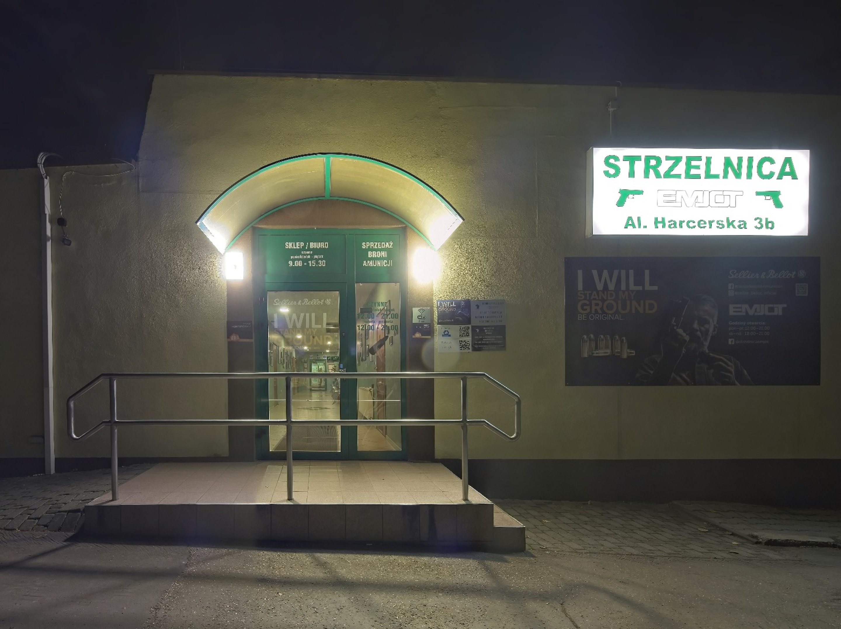 Nocne wejście do Strzelnicy EMJOT przy Al. Harcerskiej 3b. Podświetlony szyld i drzwi wejściowe do sklepu i biura strzelnicy.